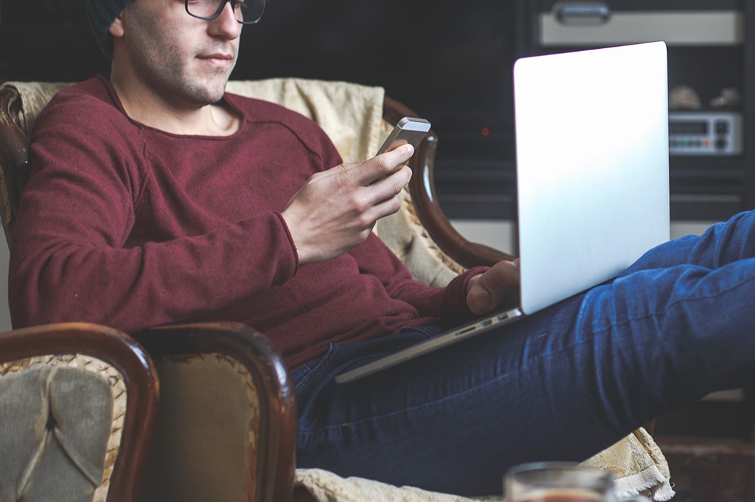 Man sitting on an armchair watching his phone and laptop Man sitting on an armchair watching his phone and laptop